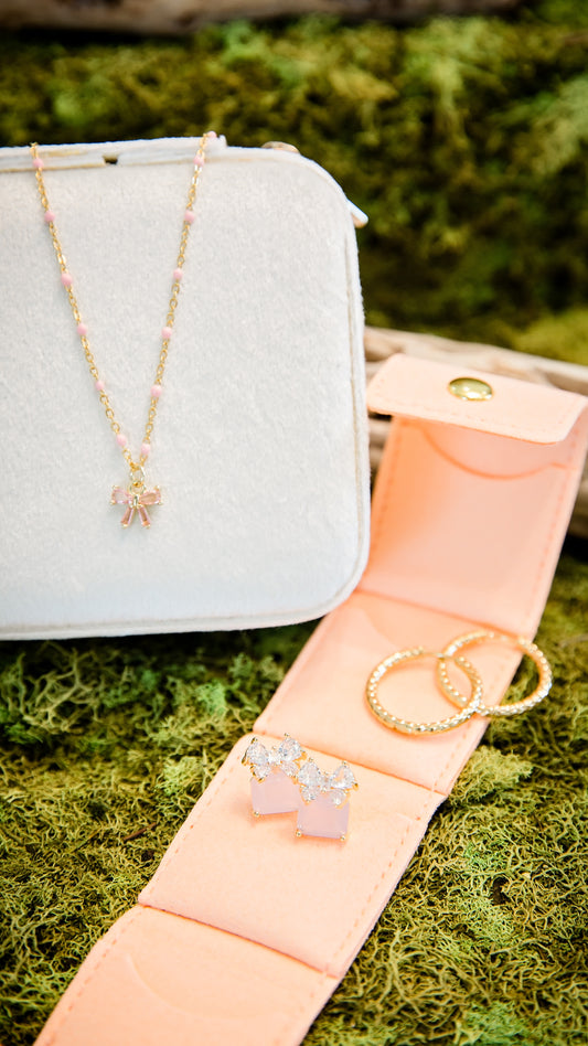 Ava Bow Necklace worn on model featuring dainty gold chain, pink bow charm, and feminine everyday jewelry styling and peach foldable jewelry travel case opened flat holding rings, studs, and delicate necklaces in soft faux suede organizer.