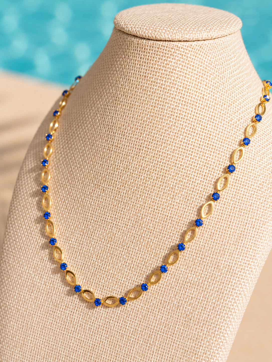 Poolside Luxe Necklace featuring genuine blue zircon stones on an 18kt gold-plated chain displayed on a jewelry bust in a poolside setting.
